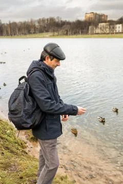 Handsome dark-haired middle-aged man in jacket, cap and backpack feeds ducks  Stock Photos