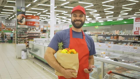 Handsome delivery man in red uniform with paper bag standing in supermarket, he Stock Footage 92546769