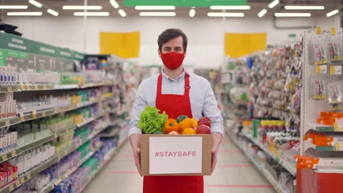 Handsome deliveryman in red face mask standing in supermarket with vegetables in Stock Footage 148069071