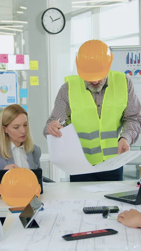 Handsome elder bearded builder in vest and helmet showing new construction bluep Stock Footage 327925535