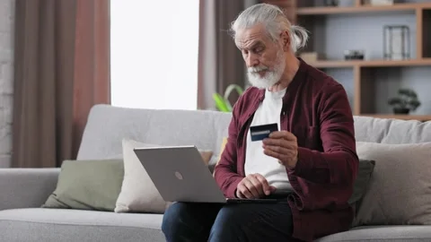 Handsome elderly gray-haired man shopping online holding credit card, sitting at Stock Footage 197413124