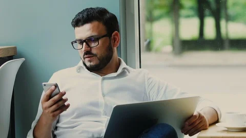 Handsome freelancer sitting near window with laptop and using his phone Stock Footage 112989397