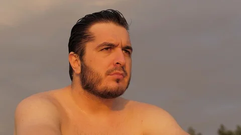 Handsome guy with bare torso and wet hair sits on beach Stock Footage 124024309