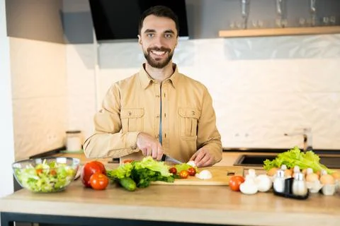 Handsome guy with beard and nice smile is cooking salad. He probably likes Stock Photos