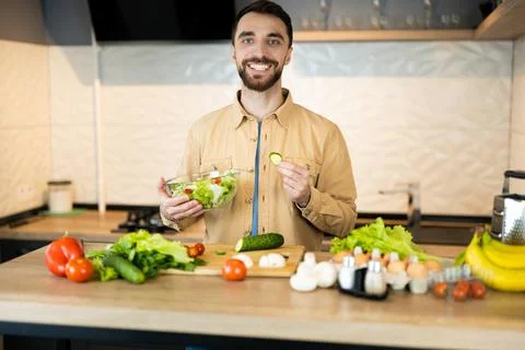 Handsome guy with beard and nice smile is cooking salad. He probably likes Stock Photos
