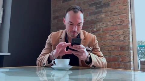 A handsome guy drinks big coffee alone in a cafe and writes on a smartphone Stock Footage 128285659