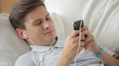 Handsome guy lying on sofa listening to music on mobile phone and smiling  Stock Footage 27303456