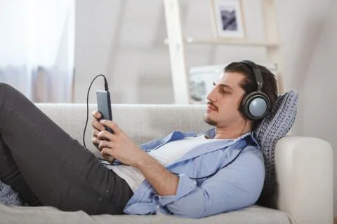 Handsome guy lying on sofa with tablet and headphones Stock Photos
