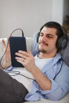 Handsome guy lying on sofa with tablet and headphones Stock Photos