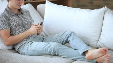 Handsome guy relaxing on sofa using cell phone loooking at camera and smiling Stock Footage 34279603