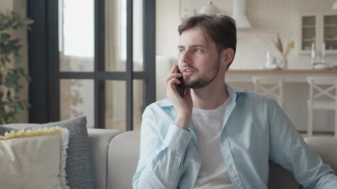 Handsome Guy Sits on Sofa and Talks on Phone in the Apartment Stock Footage 295194568