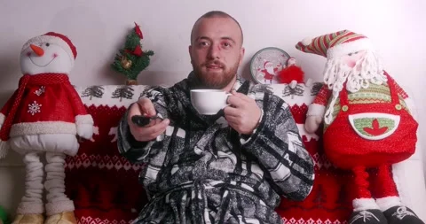 Handsome guy sitting on the sofa drinking hot tea while watching TV at Christmas Stock Footage 144355999