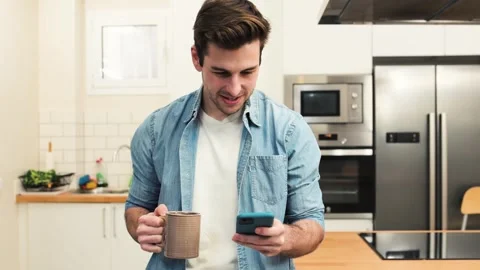 Handsome happy man looking at his phone with a cup of tea in the kitchen. young Stock Footage 248517958