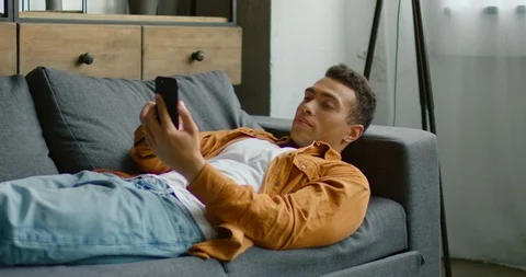 Handsome Hispanic man is relaxing on sofa, browsing in his smartphone Stock Footage 114643293