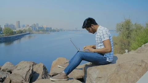 Handsome Indian Guy in Glasses is Working on a Laptop, Sitting on a Stone on the Stock Footage 111206674
