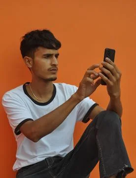 A handsome Indian guy using his phone while sitting against orange wall  Stock Photos