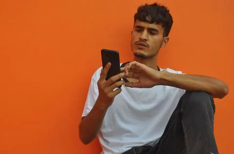 A handsome Indian guy using his phone while sitting against orange wall  Foto stock