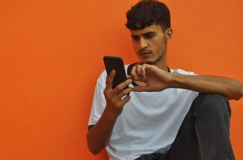 A handsome Indian guy using his phone while sitting against orange wall  Stock Photos
