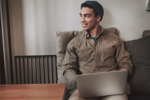 Handsome joyful guy in hat using modern silver laptop Stock Photos