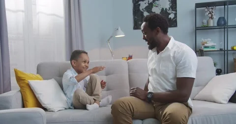 Handsome joyful man having fun with small son and smiling in living room at Stock Footage 137153127
