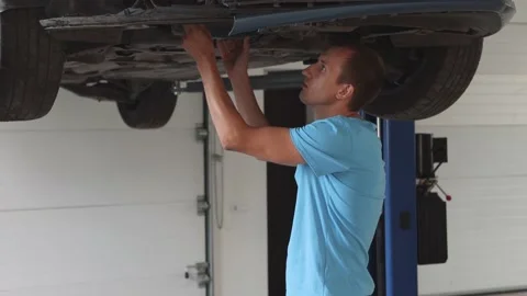 A handsome-looking young man in blue clothes inspects a car lifted Stock Footage 159220885