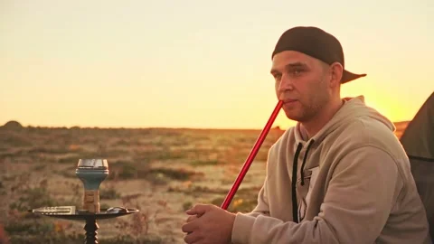 Handsome man in baseball cap smokes a hookah on sandy beach against the sunset Video stock 188737814