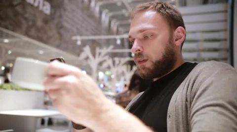 Handsome man with a beard gladly sits in a cafe, drinks coffee or tea, uses his Stock Footage 67947824