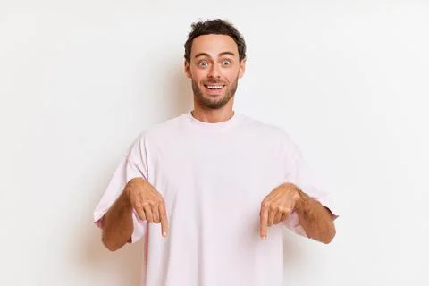 Handsome man with beard pointing down, smiles surprisedly wears white casual t Stock Photos