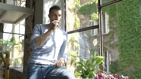 Handsome man in blue sweater eating bread next to the window and smiling to the  스톡 동영상 80257582
