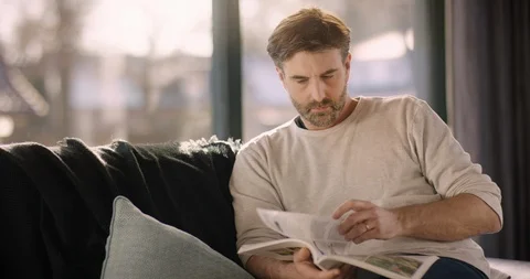Handsome Man browsing through magazine while sitting on Couch Stock Footage 86483200