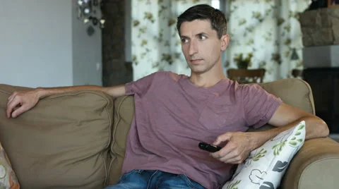 Handsome man changing TV channels with remote control sitting on sofa at home Stock Footage 65992721