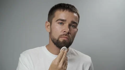 Handsome man is cleaning his face with a cotton pad on gray background Stock Footage 233517880