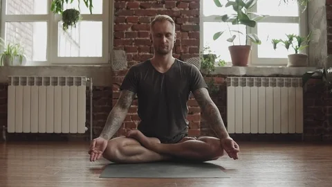 Handsome man doing yoga in lotus pose in studio with wooden floor and windows. Video stock 112734652