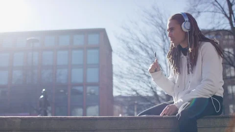 Handsome man with dreadlocks writing and listening to music as he sits on a wall Stock Footage 103806304