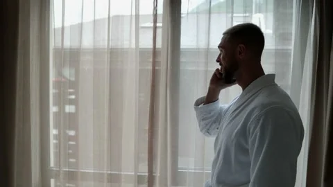 Handsome man with e-cigarette and mobile phone is standing near window in hotel Vídeos de archivo 163363759