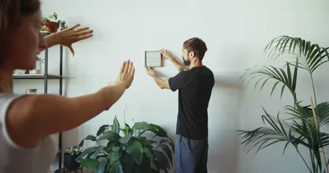 Handsome man finding place on wall to hang handmade photo frame. Young happy Stock Footage 139571661
