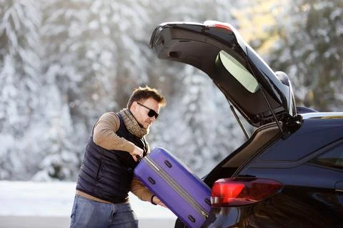 Handsome man going to vacations, loading his suitcase in car trunk. Stock Photos