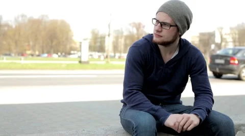 Handsome man in grey hat and glasses sitting near the street and looking around. Stock Footage 49536337