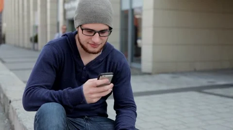 Handsome man in grey hat and glasses sitting near building and writing message Stock Footage 49537485