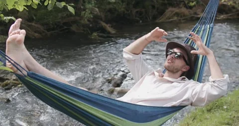 Handsome man in a hat and sunglasses lying and swinging in a hammock on a summer Stock Footage 135456068