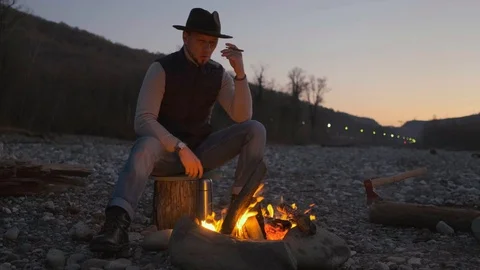 Handsome man in a hat at night sitting on a log near the fire Smoking a Stock Footage 123301302