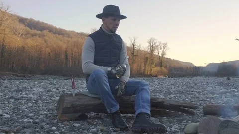 Handsome man in a hat sitting on a log near a fire drinking beer and enjoying Stock Footage 123302351
