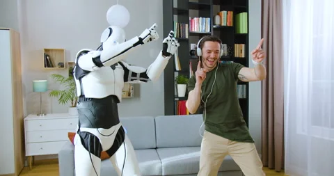 Handsome man in headphones dancing with bionic automated cyborg in living room Stock Footage 244138480
