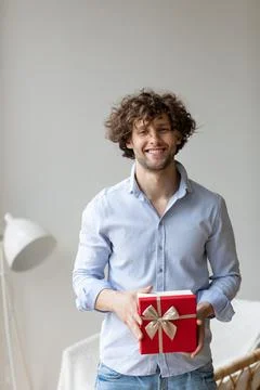 Handsome man holding a gift in a blue shirt with curly hair at home. Stock Photos