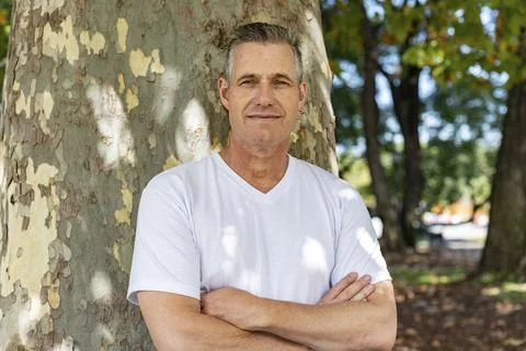 A handsome man leaning on a tree with his arms folded and looking at the came Stock Photos