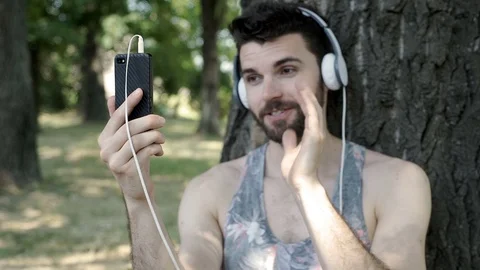 Handsome man leaning on the tree in the park and having a videocall on smartphon 스톡 동영상 78569879