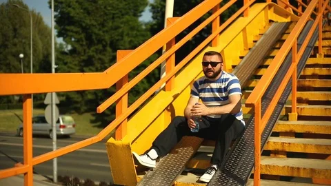 Handsome man looks tired while sitting on the stairs and drinking water Stock Footage