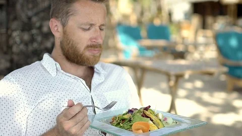 Handsome man looks unhappy while eating tasteless lunch on the tropical island,  Vídeos de archivo 80886434