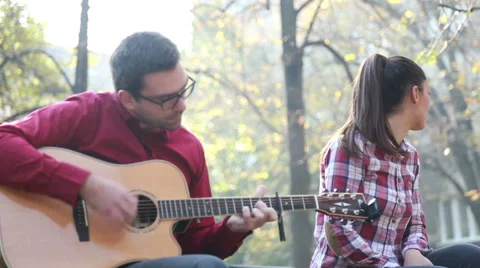 Handsome man playing guitar while beautiful woman sitting next to him in park Stock Footage 59557142