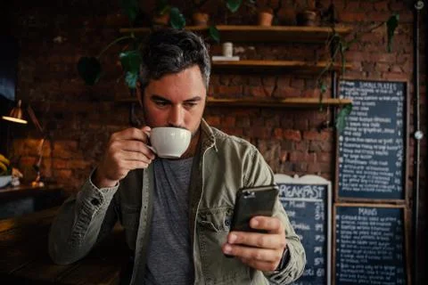 Handsome man sipping coffee while scrolling on cellular device. Stock Photos